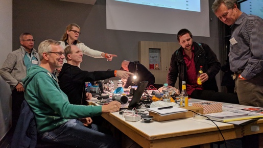 Menschen sitzen um einen Gruppentisch im Foyer des Audimax. Auf dem Tisch liegen diverse verschiedene elektronische Geräte. Mehrere Personen zeigen auf den vorderen Bereich des Tisches.